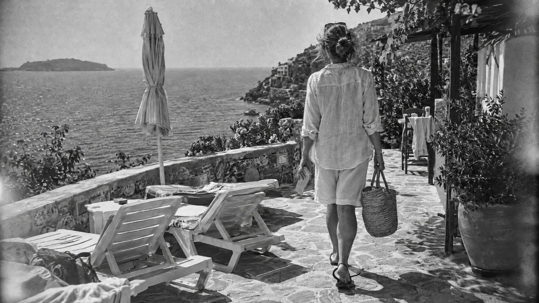 A woman in a linen shirt and shorts walks along a clifftop hotel terrace toward the sea, carrying a straw bag