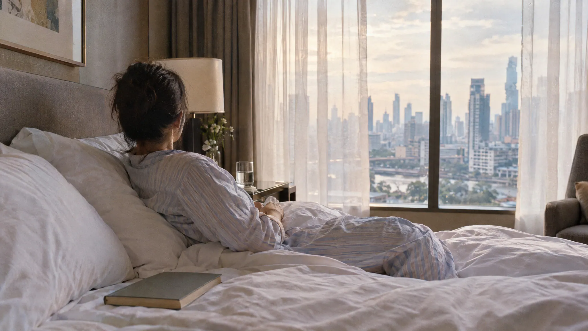 A woman resting on a hotel bed after surgery, turned toward the window and the Bangkok skyline